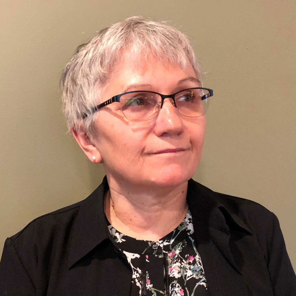 An older woman with short gray hair and glasses looks slightly to the side. She wears a black jacket over a floral top and stands in front of a plain light green wall.