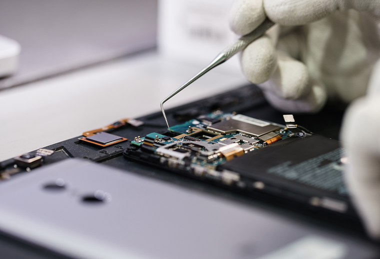 A gloved hand uses tweezers to work on the exposed circuit board of an electronic device, possibly a smartphone or tablet, highlighting detailed internal components and precise electronic repair.