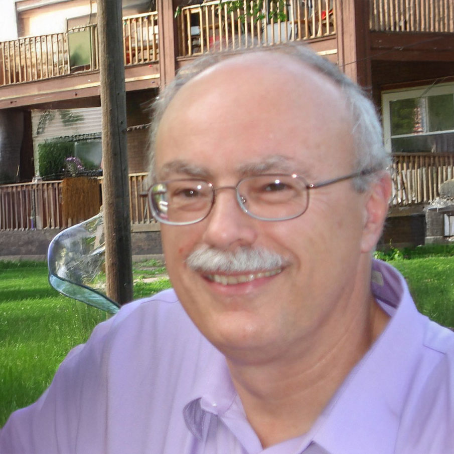 A smiling older man with glasses, a mustache, and thinning gray hair wears a light purple shirt while sitting outside. Wooden balconies and green grass are visible in the background.