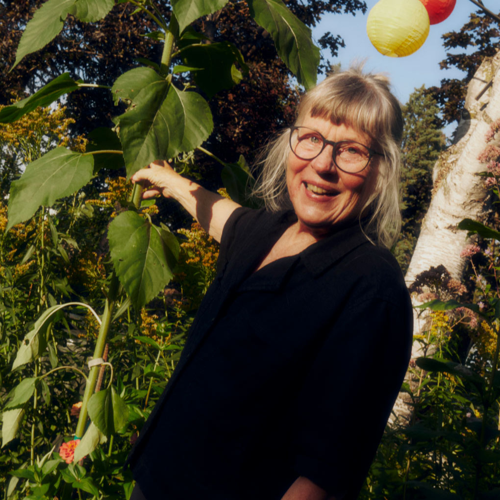 A smiling woman with glasses and blonde hair stands outdoors among tall green plants, holding a large stem. Colorful lanterns hang in the background, and sunlight creates a warm atmosphere.