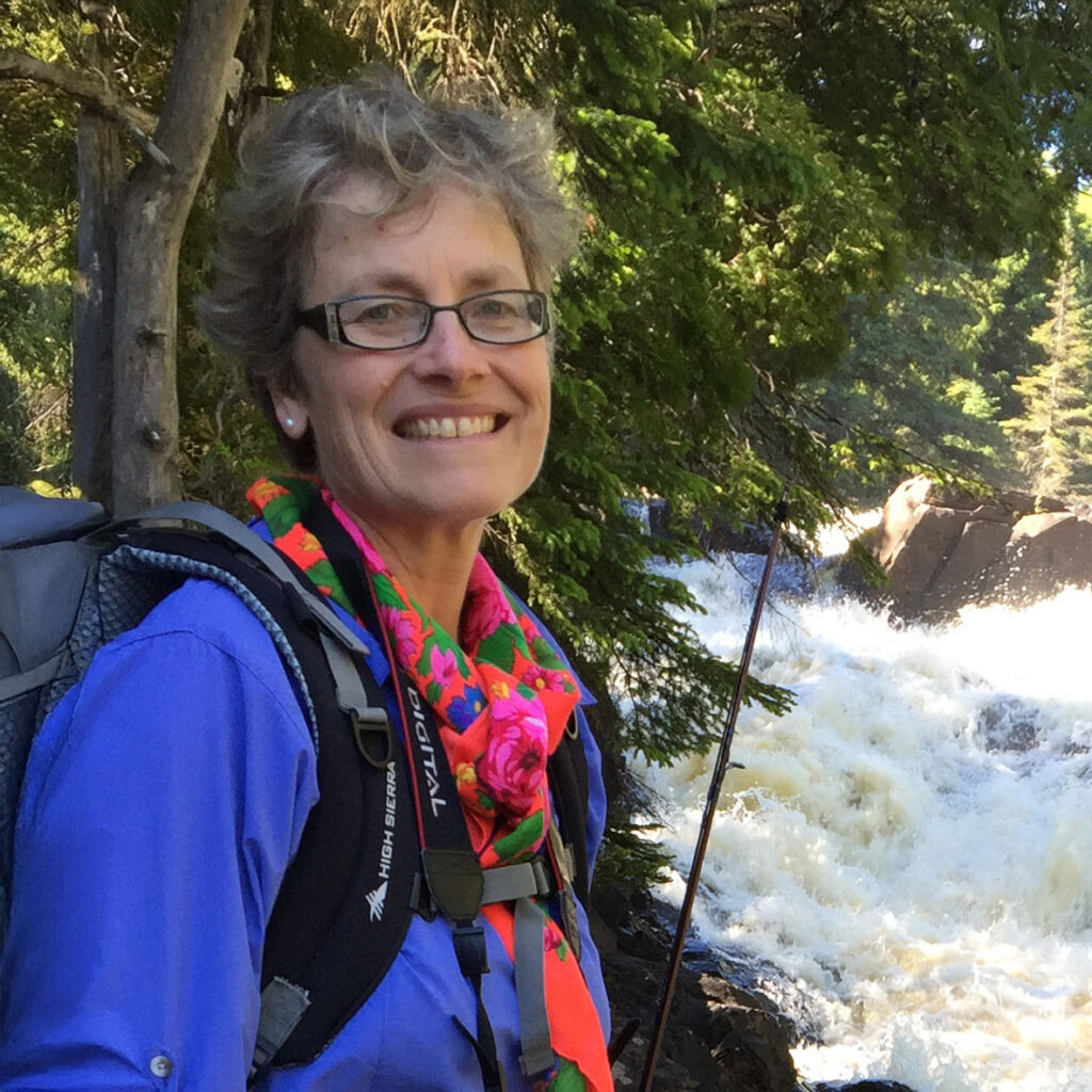 A smiling person with short gray hair, glasses, and a colorful scarf stands outdoors by a rushing river, wearing a blue shirt and backpack, surrounded by green trees on a sunny day.