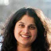 A woman with long, dark, wavy hair smiles at the camera. She is outdoors with a blurred background and is wearing a dark-colored top.