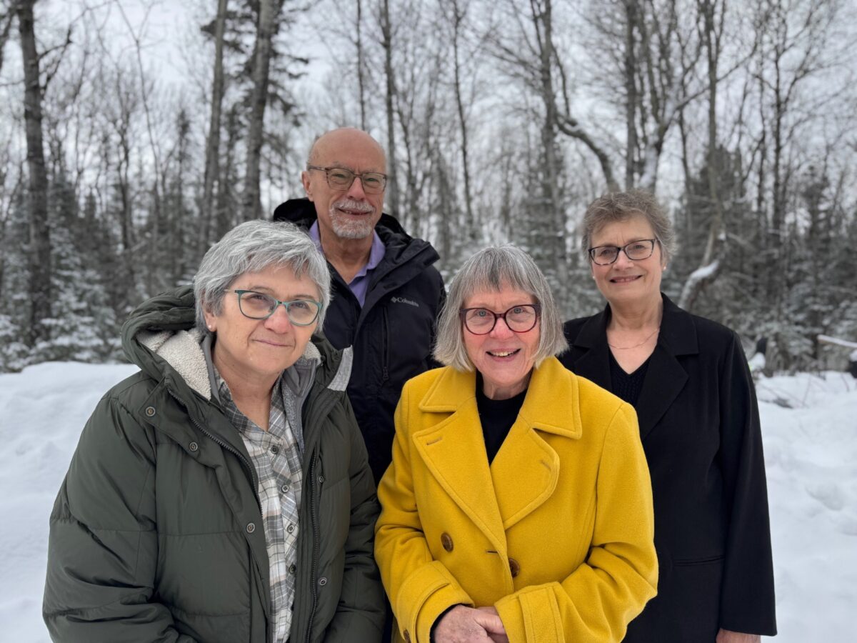 Four older adults, three women and one man, stand together outside in a snowy, wooded area. One woman wears a yellow coat, another a green coat, the man wears a black jacket, and the third woman wears a black coat. All are smiling.