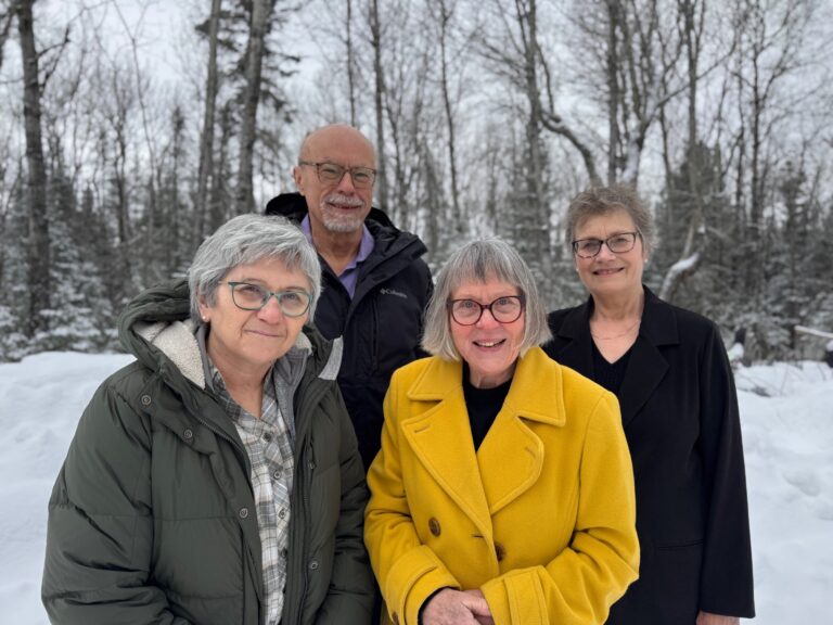 Four older adults, three women and one man, stand together outside in a snowy, wooded area. One woman wears a yellow coat, another a green coat, the man wears a black jacket, and the third woman wears a black coat. All are smiling.