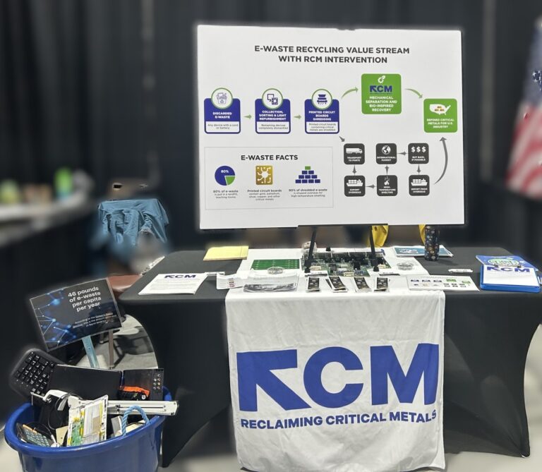 E-waste recycling display booth with an informational poster, brochures, electronic device parts on a table, and a bin filled with discarded electronics. A banner reads RCM Reclaiming Critical Metals.
